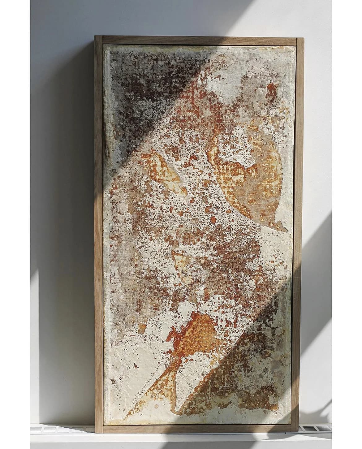 Panel in a wooden frame leaning against a wall, with a diagonal band of sunlight across its rust, cream and grey surface.