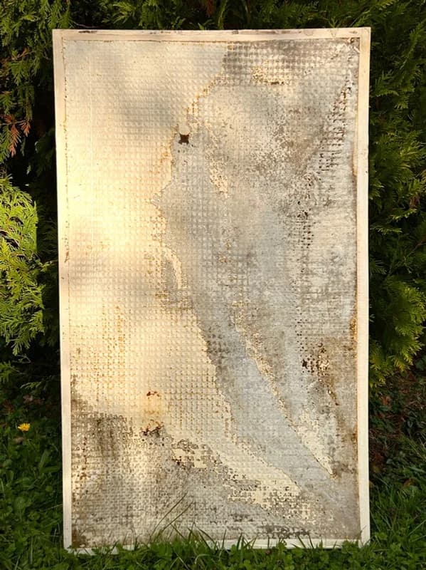 Panel with a dotted grid texture and soft grey patches, propped against a hedge in sunlight.
