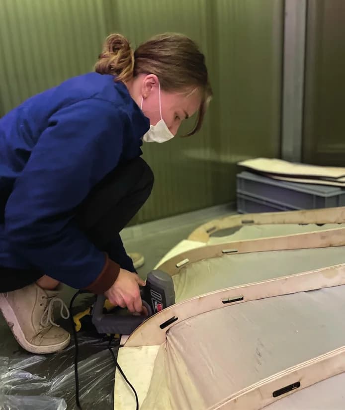 A maker kneeling on the floor, fixing pale fabric to curved plywood ribs with a hand tool.
