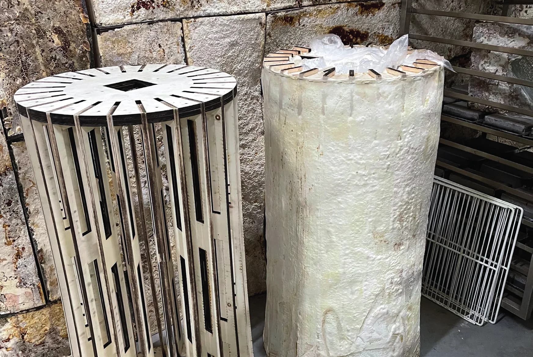 A cylindrical laser-cut wooden skeleton with a slotted top disc and vertical ribs, standing beside a grown column wrapped in its envelope.